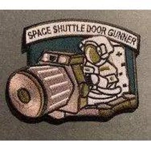 May include: An embroidered patch featuring a cartoon astronaut operating a large camera, with the text SPACE SHUTTLE DOOR GUNNER arched above in black lettering on a white background. The design includes shades of blue, white, and tan.