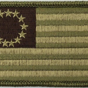 May include: Embroidered patch featuring a colonial American flag design. The patch has a dark brown canton with a circle of stars and alternating green and tan stripes. The border is green.