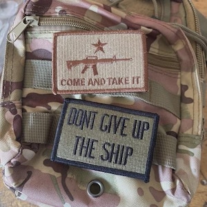 May include: A camouflage tactical bag featuring two rectangular morale patches. The top patch is tan with a brown rifle, a star, and the text COME AND TAKE IT. The bottom patch is olive green with a black border and the text DONT GIVE UP THE SHIP.