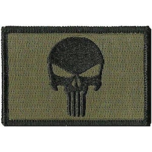 Puede incluir: Un parche rectangular bordado que presenta un fondo verde oliva con un emblema de calavera negro cosido en el centro. El parche tiene un borde negro grueso y una superficie de tela texturizada, mostrando un trabajo de hilo detallado en el diseño.