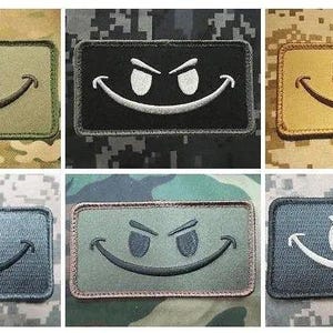May include: A collection of six rectangular embroidered patches featuring a smiling face with angry eyes. The patches come in various colors, including olive green, black, and gold, with different background patterns.