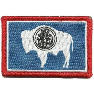 May include: A rectangular embroidered patch featuring the Wyoming state flag design. It displays a white silhouette of an American bison centered on a blue field, with a white border and a thick red stitched edge surrounding the entire rectangular shape.
