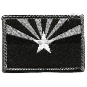 May include: A rectangular embroidered patch featuring a black and silver color scheme. The design displays a central five-pointed star with radiating sunbeams extending toward the upper corners, all bordered by a thick, dark gray stitched edge.