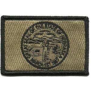 May include: A rectangular embroidered patch in shades of tan and black. The center features a circular emblem depicting a landscape with a figure, surrounded by the text THE GREAT SEAL OF THE STATE OF NEBRASKA MARCH 1ST 1867 in a black border.