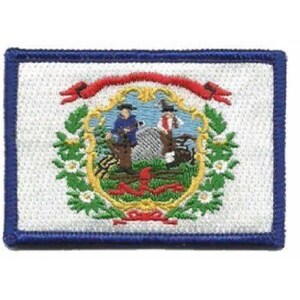 May include: A rectangular embroidered patch featuring the West Virginia state flag design. The white background is bordered in blue, with a central emblem showing two figures, a plow, and a rock, surrounded by a wreath of rhododendron flowers.