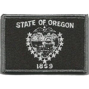 Peut inclure: Un écusson rectangulaire brodé noir avec le texte STATE OF OREGON en blanc en haut et 1859 en bas. Le centre présente un emblème brodé blanc du sceau de l'État entouré d'un cercle de petites étoiles sur un fond en tissu noir.