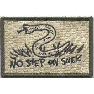Pode incluir: Um emblema retangular bordado com uma borda verde-oliva e fundo bege. O design apresenta um gráfico bordado em marrom escuro de uma cobra enrolada com língua, posicionado acima do texto NO STEP ON SNEK em uma fonte negrito marrom escuro.