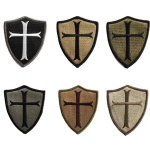May include: Six shield-shaped patches with a cross design. The patches are in various colors, including black, tan, and olive green. The crosses are in contrasting colors, such as white and black. The patches are likely for clothing or accessories.