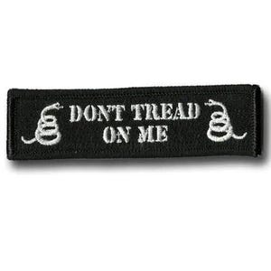 Puede incluir: Un parche rectangular bordado en negro con texto blanco que dice DONT TREAD ON ME. El parche incluye un gráfico de una serpiente enroscada blanca a la izquierda y a la derecha del texto, todo bordeado por un grueso borde cosido en negro.