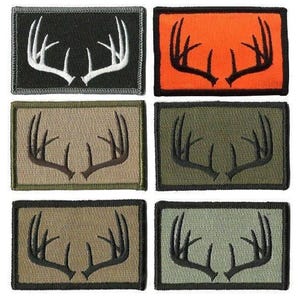 May include: Rectangular patches with embroidered antler designs. The patches come in various colors, including black with white antlers, orange with black antlers, and shades of green and tan with brown or black antlers. Each patch has a contrasting border.