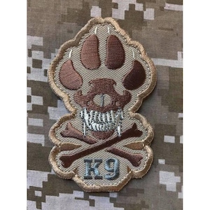 May include: A tan and brown embroidered patch featuring a stylized paw print design with a skull and crossbones motif in the center. The text K9 is stitched in light blue at the bottom of the patch, which has a tan border and a jagged edge.