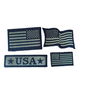 May include: Four embroidered patches featuring the United States flag in a monochromatic dark blue and light gray color scheme. One rectangular patch displays the text USA flanked by two stars, while the others show flat or waving flag designs.