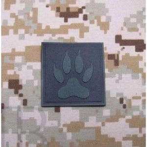 Puede incluir: Un parche cuadrado de color gris oscuro que presenta un diseño de huella de pata en relieve en el centro. El parche tiene un acabado mate suave y un borde definido, mostrando un gráfico claro y único de huella animal sobre un fondo gris.