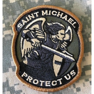 Pode incluir: Um emblema bordado circular com a representação de São Miguel com asas e uma espada. O texto SAINT MICHAEL está arqueado no topo e PROTECT US na parte inferior em fio branco sobre um fundo escuro, com uma borda de cor dourada.