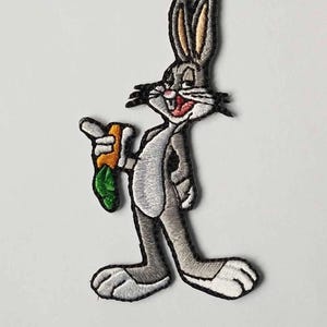 May include: Embroidered patch featuring Bugs Bunny, the iconic cartoon character. Bugs is depicted in a standing pose, holding a carrot. The patch is primarily gray, with white, orange, and green accents, and a black outline.