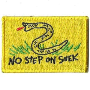Pode incluir: Um emblema retangular bordado em amarelo com uma cobra preta de língua vermelha, lâminas de grama verde e o texto preto NO STEP ON SNEK. O design é contornado por uma borda costurada em amarelo, criando um emblema gráfico detalhado.