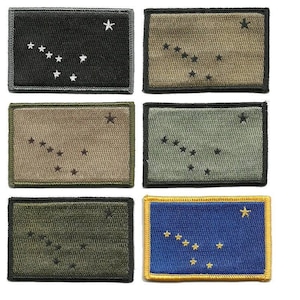 May include: Rectangular patches featuring the Alaska state flag design. The patches come in various colors, including black, olive green, tan, and blue. Each patch has a black border and embroidered stars representing the Big Dipper and the North Star.