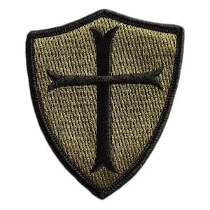 May include: An embroidered patch in the shape of a shield with a pointed bottom. The patch features an olive green background with a black border and a prominent black cross centered on the shield, showcasing detailed thread texture throughout.