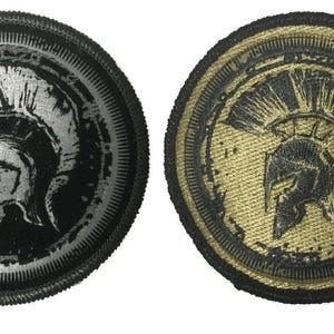 May include: Two round embroidered patches featuring a Spartan helmet design. One patch is black with a silver helmet, the other is gold with a black helmet. Both have a black border and detailed stitching.