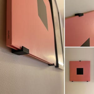 May include: A square pink artwork featuring a black square and a grey triangle design. The artwork is mounted on a white wall with black mounting hardware. The text "JAG450" is visible on the side of the artwork.