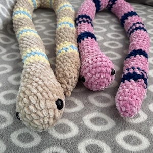 May include: Two plush snake toys made of soft, textured yarn. One snake is beige with yellow and light blue stripes, while the other is pink with dark blue stripes. Both snakes feature small, round, black plastic eyes on their rounded heads.