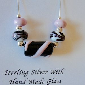 May include: A handmade glass necklace with a black rectangular center bead and pink swirl design. The necklace features round, patterned beads in white, black, and pink, and small silver beads. The text "Sterling Silver With Hand Made Glass" is below the necklace.