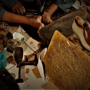 May include: A cobbler's workshop with tools and materials. Includes shoe lasts, a leather cutting board, a metal shoe form, and a can of adhesive. The cobbler is working on a shoe, using a hammer.