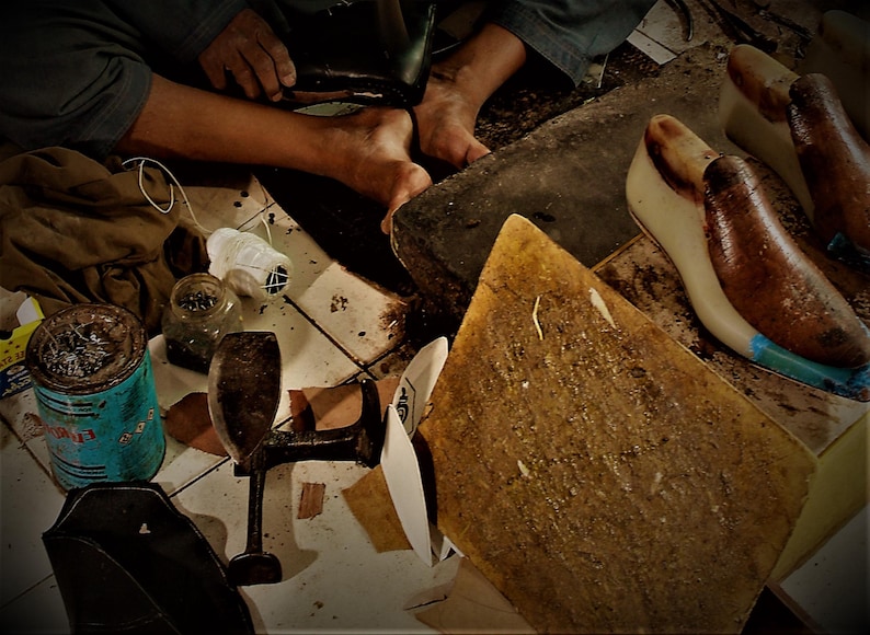 May include: A cobbler's workshop with tools and materials. Includes shoe lasts, a leather cutting board, a metal shoe form, and a can of adhesive. The cobbler is working on a shoe, using a hammer.