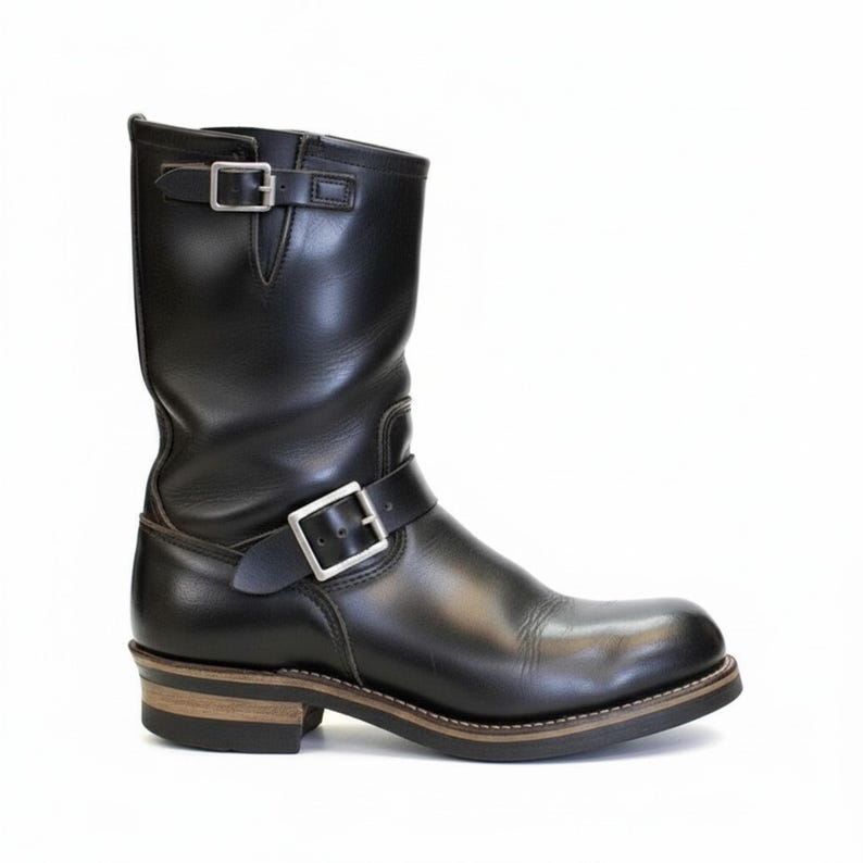 May include: A black leather motorcycle boot with a mid-calf height. The boot features a rounded toe, a low heel, and two adjustable straps with silver buckles. The sole has a brown and black layered design.