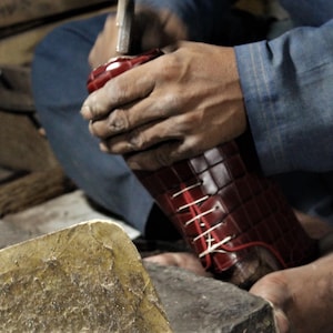 May include: A shoemaker is working on a burgundy leather shoe. The shoe is being hammered, with white laces visible. The shoe has a textured, patterned design. The shoemaker is wearing a blue shirt.