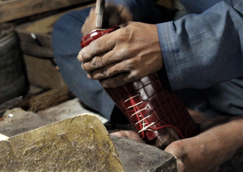 May include: A shoemaker is working on a burgundy leather shoe. The shoe is being hammered, with white laces visible. The shoe has a textured, patterned design. The shoemaker is wearing a blue shirt.