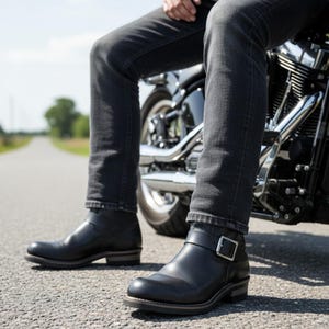 May include: Black leather motorcycle boots with a buckle fastening. The boots have a low heel and are worn with dark denim jeans. The image is taken outdoors on a paved road, with a motorcycle in the background.