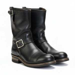 May include: A pair of black leather motorcycle boots. The boots are mid-calf length, with a rounded toe and a low heel. Each boot has a buckle strap around the ankle and another near the top. The soles have a brown and tan layered design.