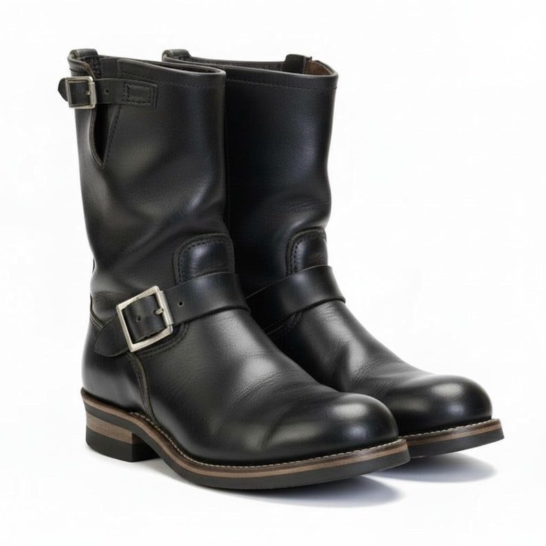 May include: A pair of black leather motorcycle boots. The boots are mid-calf length, with a rounded toe and a low heel. Each boot has a buckle strap around the ankle and another near the top. The soles have a brown and tan layered design.