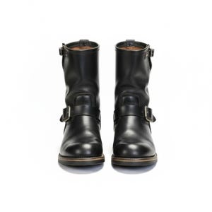 May include: Two black leather motorcycle boots with buckle straps. The boots have a rounded toe and a low heel. Each boot features a strap around the ankle and another across the foot. The background is white.