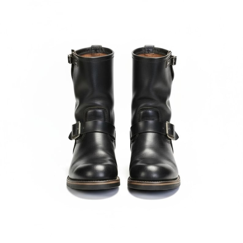 May include: Two black leather motorcycle boots with buckle straps. The boots have a rounded toe and a low heel. Each boot features a strap around the ankle and another across the foot. The background is white.