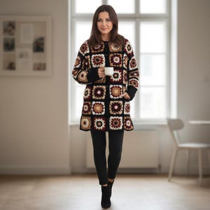 May include: A woman wearing a long, multi-colored crochet coat with a square pattern in shades of brown, beige, and burgundy. The coat has black trim and buttons. She is holding a white mug and wearing black pants and boots.