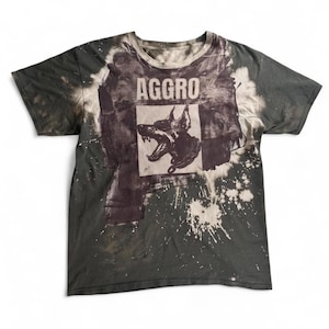 Puede incluir: Una camiseta de manga corta en gris carbón oscuro con un efecto de lavado desgastado y blanqueado. El centro presenta un gráfico rectangular blanco con la palabra AGGRO en letras negritas sobre una ilustración en blanco y negro de un Doberman.