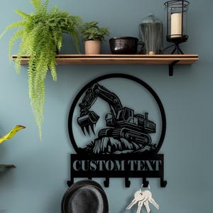 Peut inclure: Porte-clés en métal noir avec texte "CUSTOM TEXT". Le porte-clés présente un design circulaire avec une silhouette d'excavatrice. En dessous se trouvent des crochets pour clés et un chapeau. Le porte-clés est fixé sur un mur bleu.