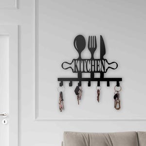 May include: Black metal kitchen-themed key holder with a spoon, fork, and knife design above the word "KITCHEN". The key holder has several hooks with keys hanging from them. The key holder is mounted on a white wall.