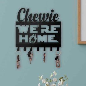 Pode incluir: Porta-chaves de metal preto com as palavras "Chewie WE'RE HOME" recortadas. O porta-chaves tem seis ganchos com chaves penduradas. Uma imagem emoldurada está pendurada na parede à direita. Uma planta com flores brancas está por baixo.
