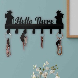 Puede incluir: Llavero de metal negro con el texto "Hello There" y dos siluetas. Varias llaves cuelgan de los ganchos. El llavero está montado en una pared azul claro.