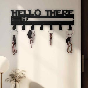 May include: Black metal key holder with the words "HELLO THERE" cut out at the top. The key holder has six hooks with keys hanging from them. A level is cut out below the words. The key holder is mounted on a white wall.