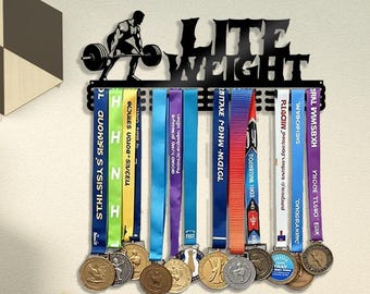 May include: Black metal medal display with the words "LITE WEIGHT" and a weightlifter silhouette. Colorful ribbons hold various medals. The display is mounted on a light-colored wall.