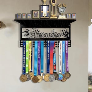 May include: Black metal medal display with the name "Alexandra" and a gymnast silhouette. The display holds medals with colorful ribbons and has a shelf for trophies and small framed pictures. A wall-mounted design.