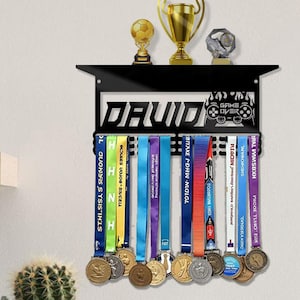 May include: Black metal medal display with the name "DAVID" cut out. The display has a top shelf holding three trophies and a "GAME OVER" design. Colorful ribbons hold various medals.