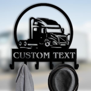 May include: Black metal wall-mounted coat rack featuring a semi-truck silhouette within a circular frame. The rack includes hooks below a panel with the text "CUSTOM TEXT". A gray hoodie and a gray hat hang from the hooks.