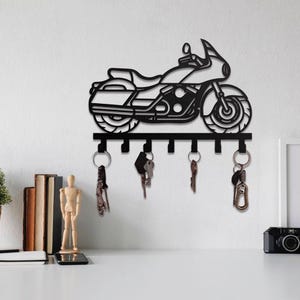 Könnte beinhalten: Schwarzer Motorrad-Schlüsselhalter aus Metall mit sechs Haken. Der Schlüsselhalter zeigt eine detaillierte Umrisszeichnung eines Motorrads, einschließlich Räder, Sitz und Lenker. Mehrere Schlüssel hängen an den Haken. Der Schlüsselhalter ist an einer weißen Wand befestigt.