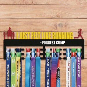 Peut inclure: Présentoir de médailles en métal noir avec la citation "I JUST FELT LIKE RUNNING - FORREST GUMP" en texte jaune. Le présentoir contient plusieurs rubans colorés, chacun avec du texte. Une silhouette d'une personne assise sur un banc et un coureur sont sur le présentoir.