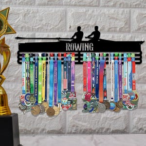 Puede incluir: Expositor de medallas de metal negro con la palabra "ROWING" y siluetas de remeros. Cintas y medallas de colores cuelgan del expositor, mostrando varios logros de carreras. El expositor está montado en una pared de ladrillos blancos.